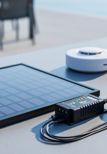 Close-up of a compact 9-volt solar panel kit on a workbench next to a smoke detector and small electronics under natural light.